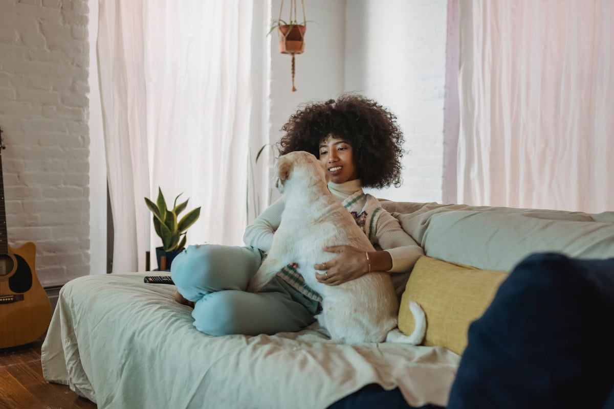 A woman petting a light-colored dog while sitting on a sofa at home.